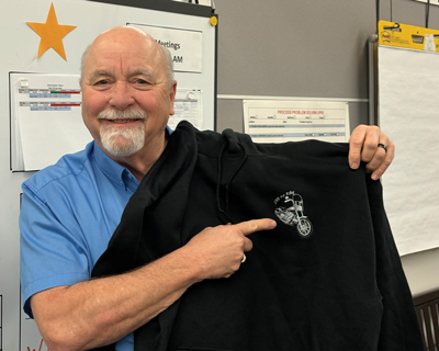 Kevin Peterson smiling while holding up and pointing to his gifted black hoodie, which is embroidered with a picture of his motorcycle